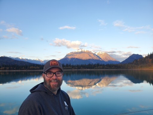 Dennis Fowler beside Lake Clark with mountains reflected in the water
