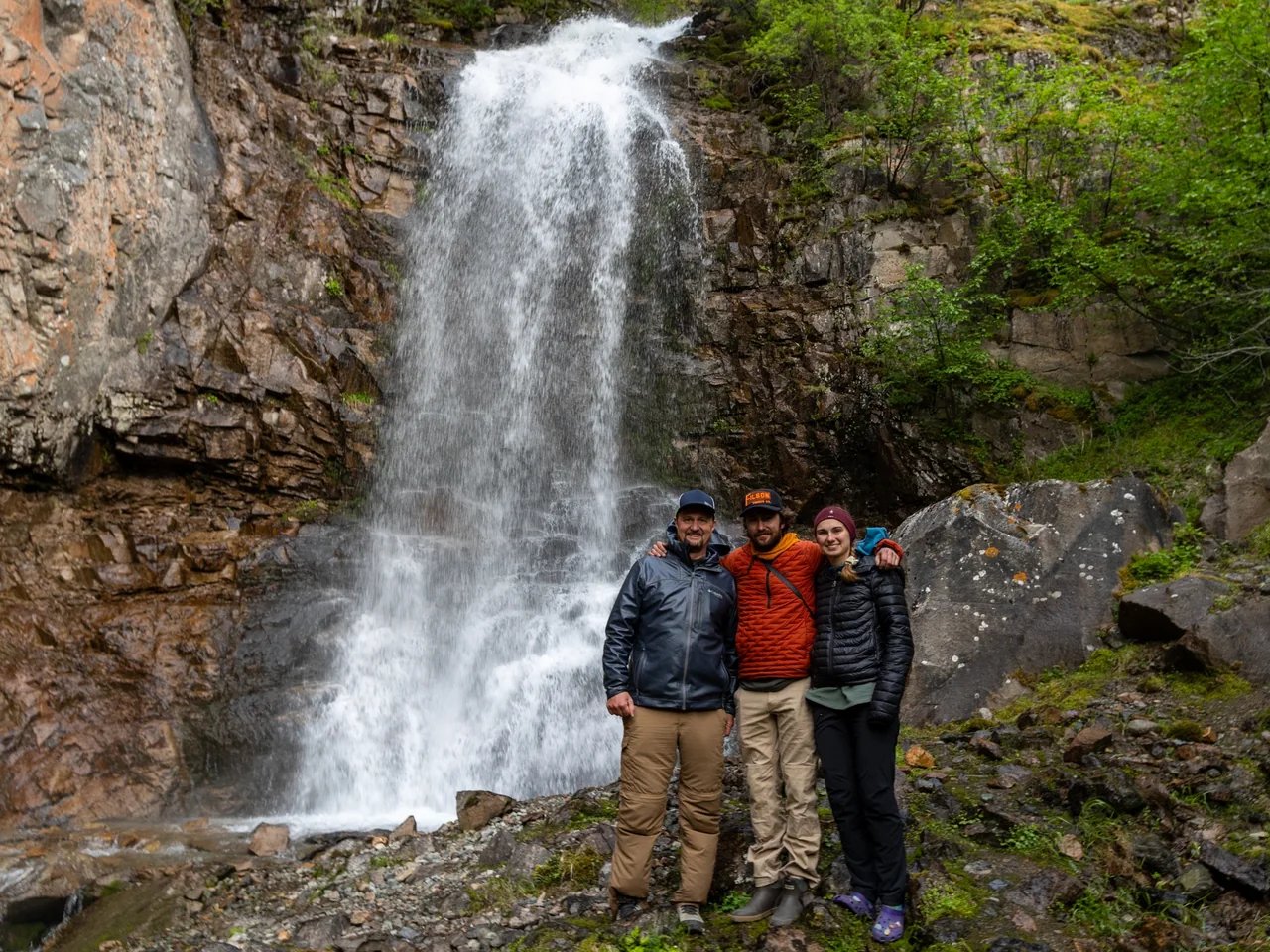 Three hikers smiling in front of a hidden waterfall during an off-boat adventure near Lake Clark