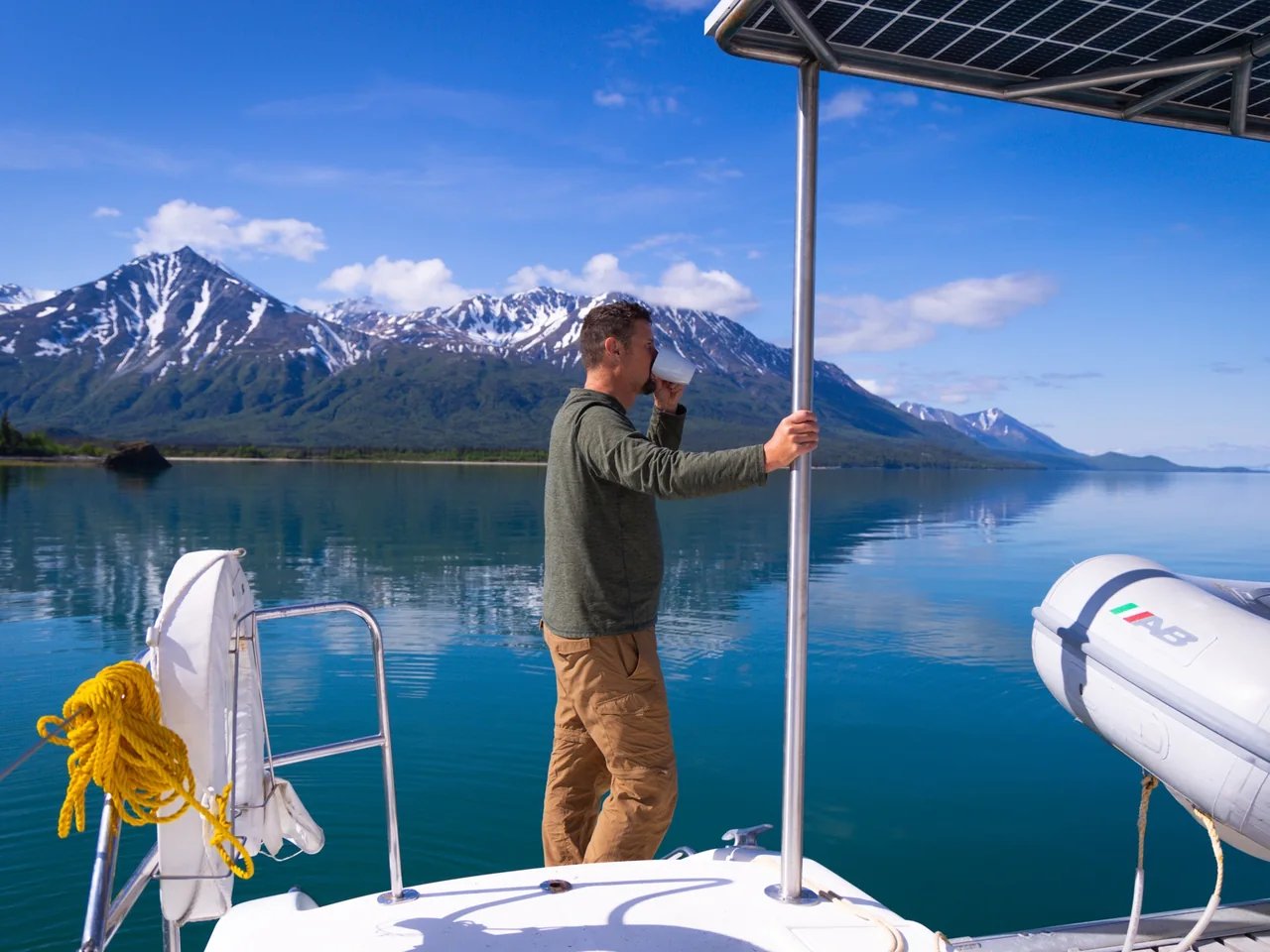 Guest sipping coffee on the deck of Odyssey with snow-covered peaks reflected in the calm waters of Lake Clark