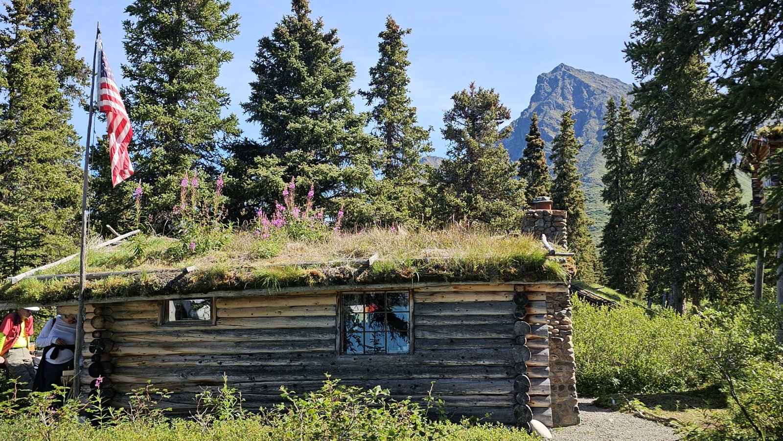 Dick Proenneke’s log cabin at Twin Lakes, with a sod roof and an American flag